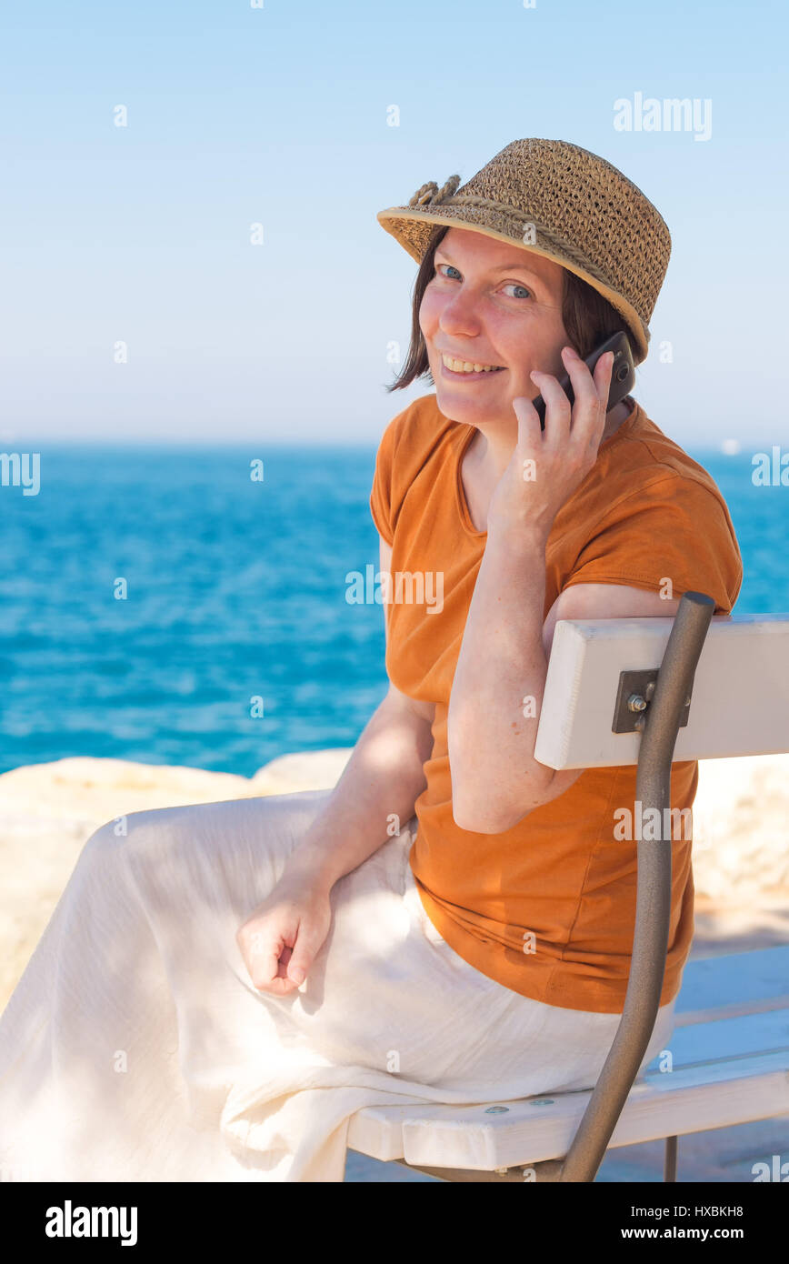 Schöne Erwachsene kaukasischen Frau tragen stilvolle Strohhut ist Mobiltelefon in den Sommerferien Urlaub am Meer Stockfoto