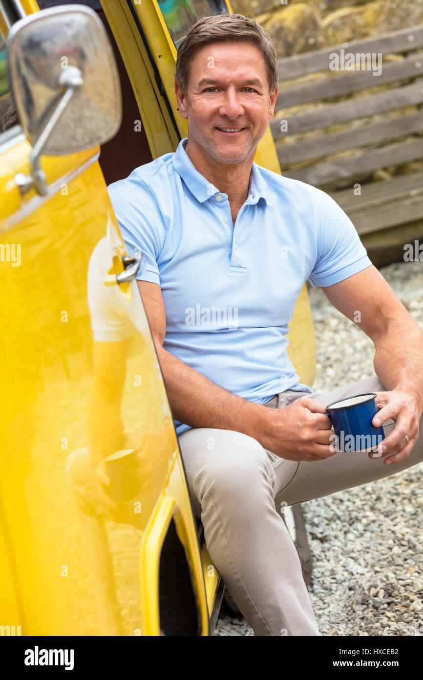 Schön, erfolgreich und glücklich Mann mittleren Alters sitzen in der Tür eines gelben Camper van Bus trinken Tee oder Kaffee Form einer Zinn Tasse oder Becher Stockfoto