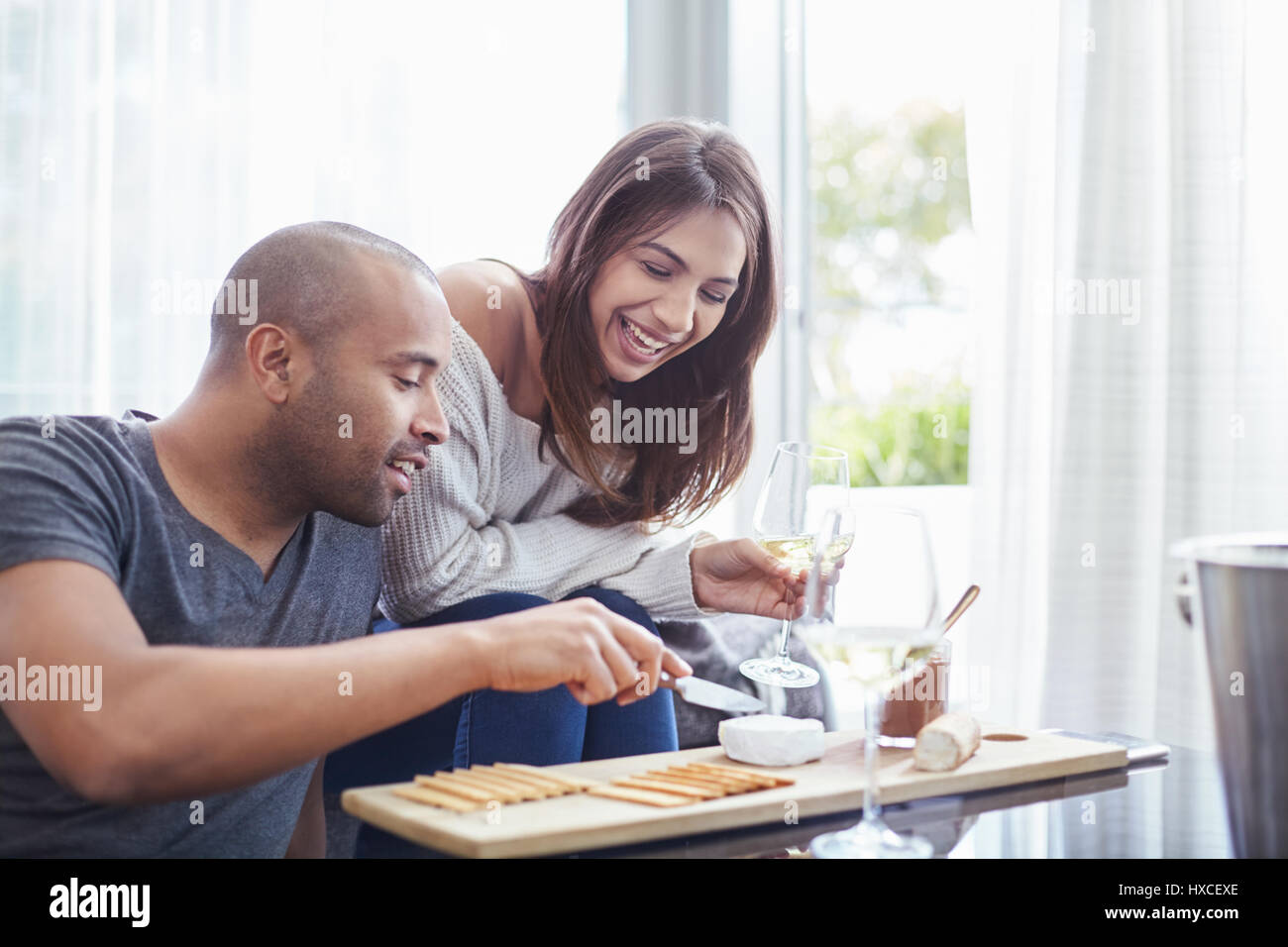 Lächelndes Paar Weißwein trinken und essen Kekse und Käse im Wohnzimmer Stockfoto
