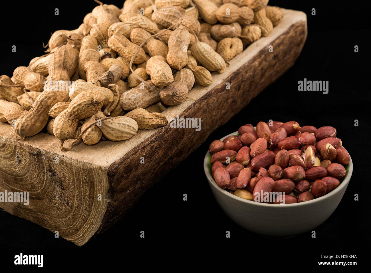 Erdnüsse in aller Kürze auf ein Stück Holz und geschälte Erdnüsse in Schüssel weiß auf schwarzem Hintergrund Stockfoto