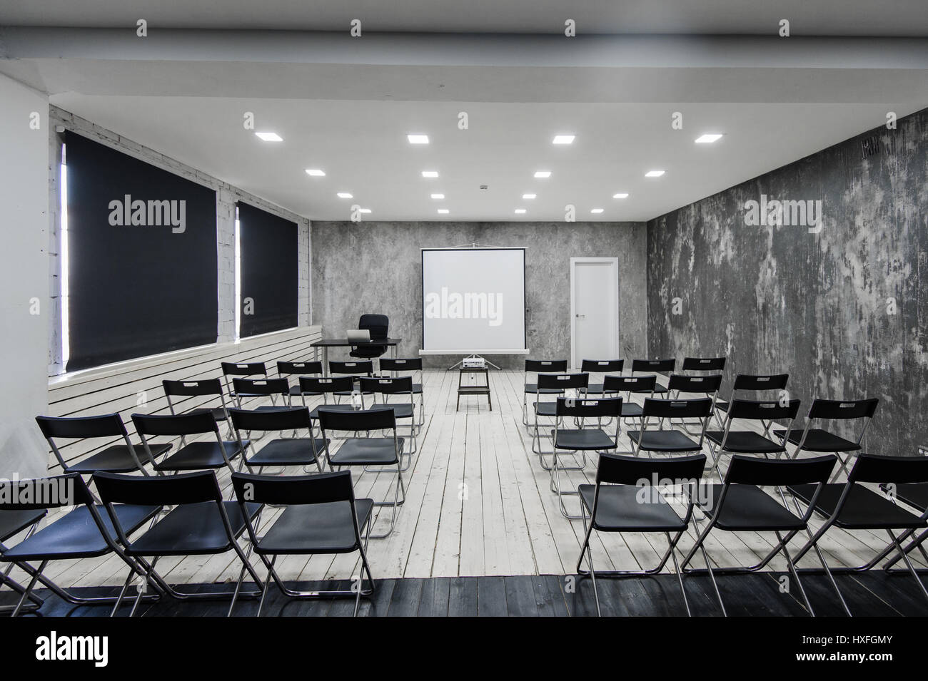 Raum für Vortrag mit vielen dunklen Stühlen. Wände sind weiß, Loft Interieur. Auf der rechten Seite ist eine Tür. Auf dem Hintergrund gibt es einen Tisch mit einem Laptop. Stockfoto