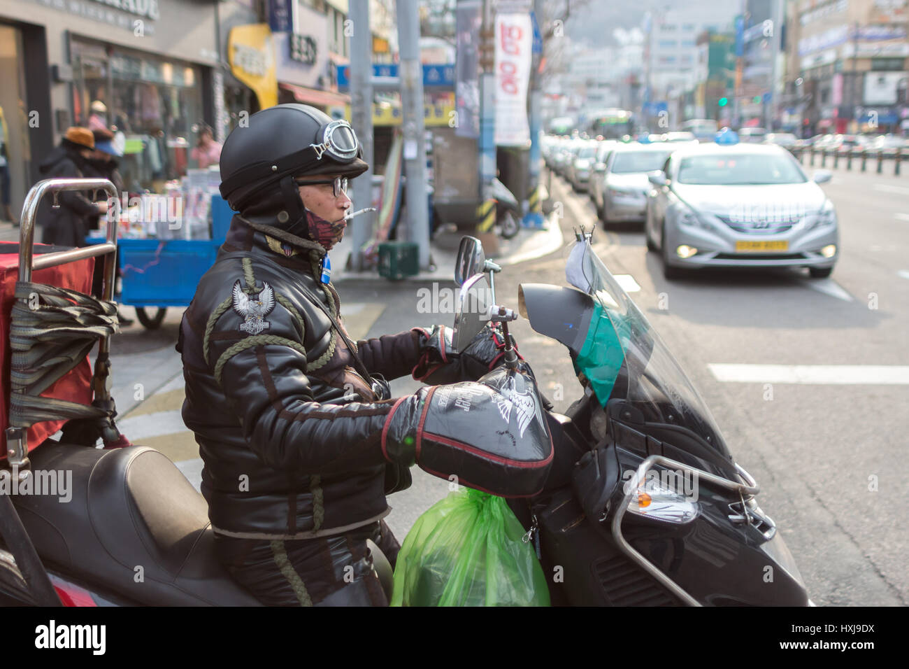 Ein Mann mit einer Zigarette auf dem Mund, der in Busan, Südkorea, ein Motorroller-Motorrad mit einer Harley Davidson Lederjacke fährt. Stockfoto