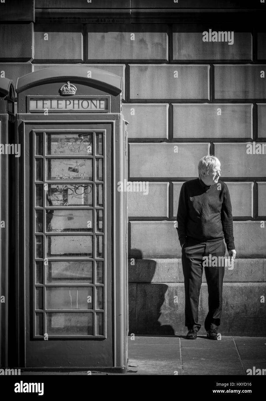 Reifer kaukasischer Mann, der neben einem traditionellen Telefonkasten steht und eine Zigarette raucht. Edinburgh. VEREINIGTES KÖNIGREICH Stockfoto