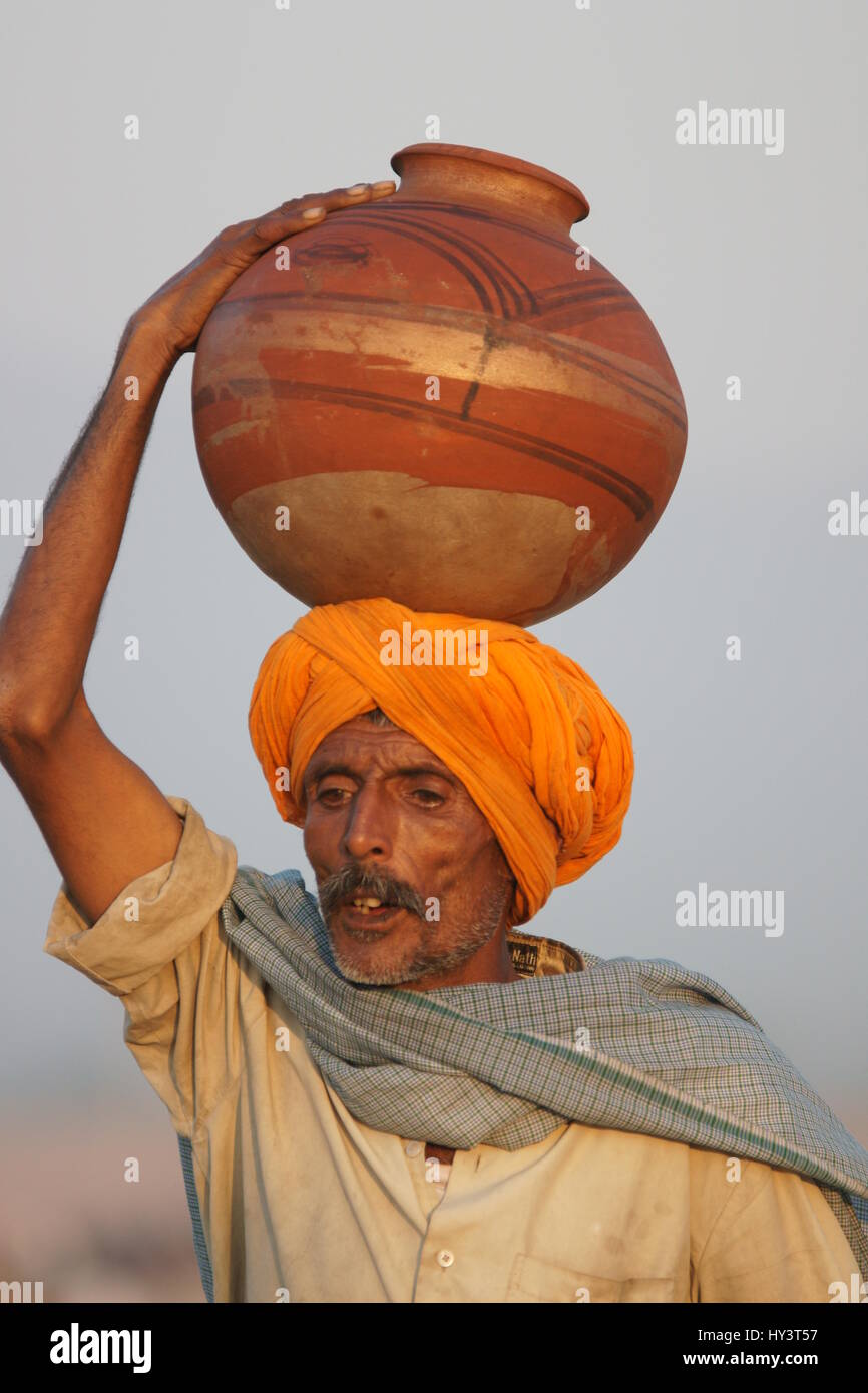 Indischer Mann in orange Turban tragen Topf mit Wasser auf dem Kopf auf der Messe Pushkar in Rajasthan, Indien Stockfoto