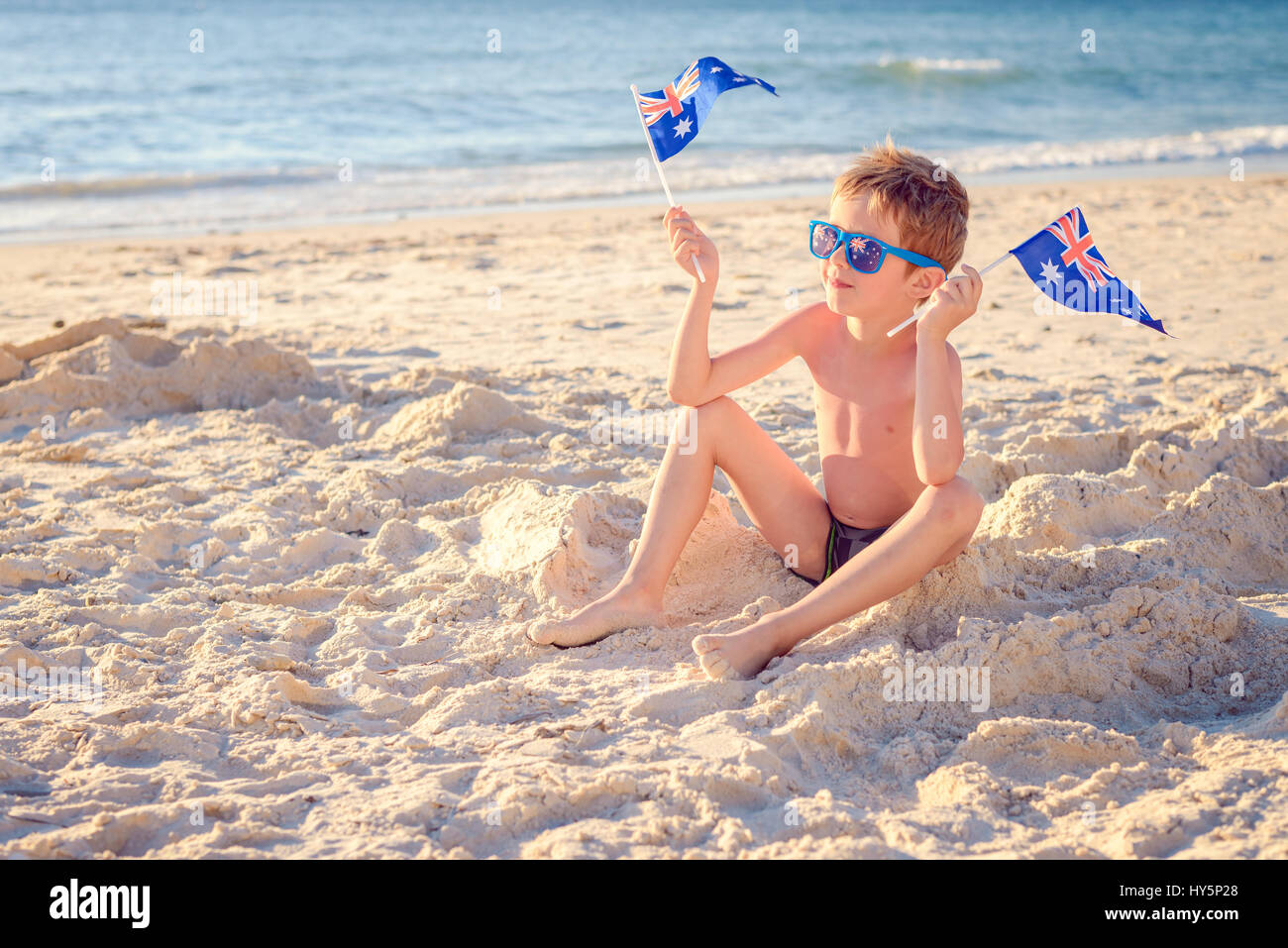 Niedlichen lächelnden jungen am Strand sitzen und halten australische Flaggen am Australia day Stockfoto