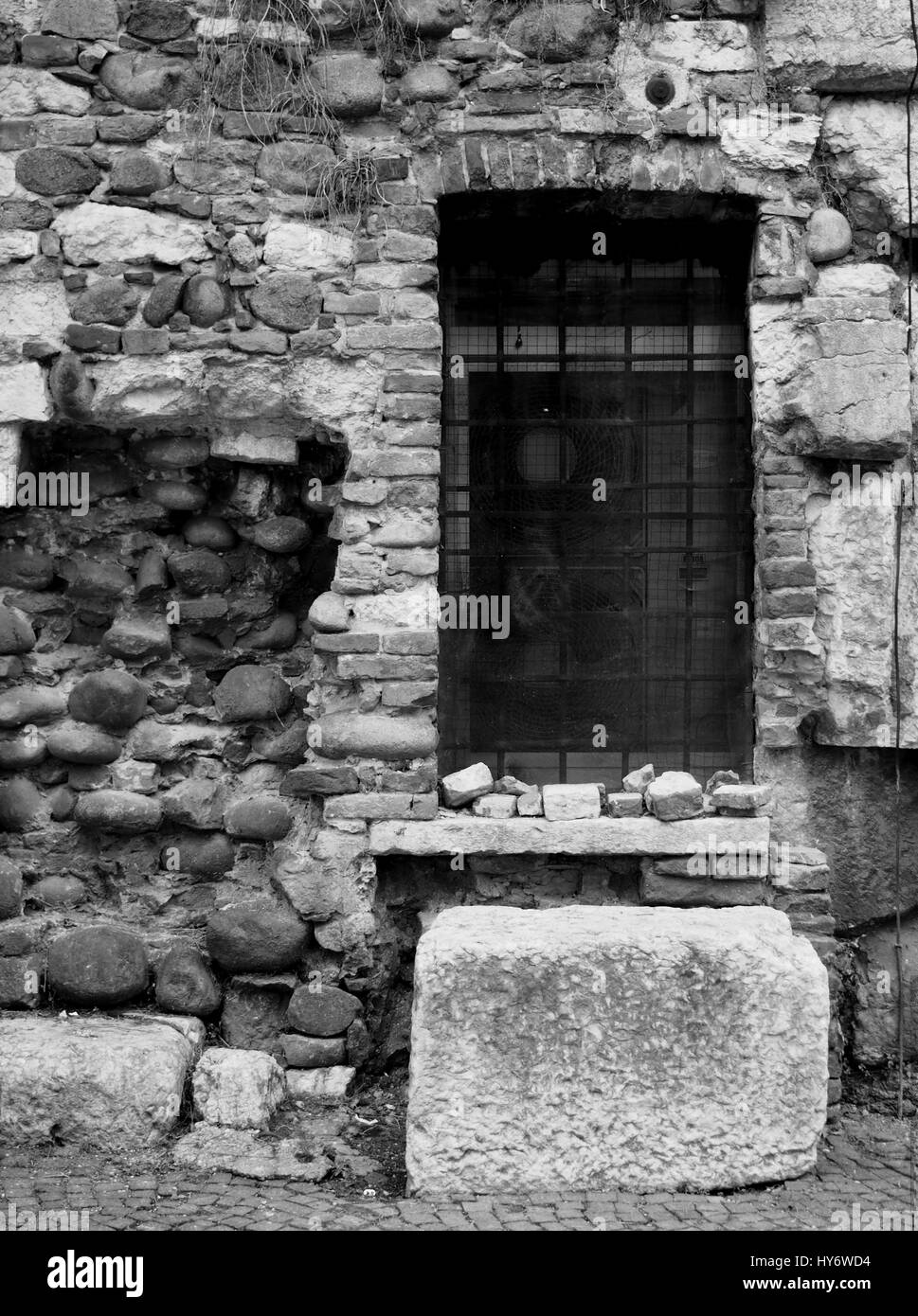 Oldamaged und reparierte Ziegelwand mit einem vergitterten Fenster und alten Steinen in Monochrom Stockfoto