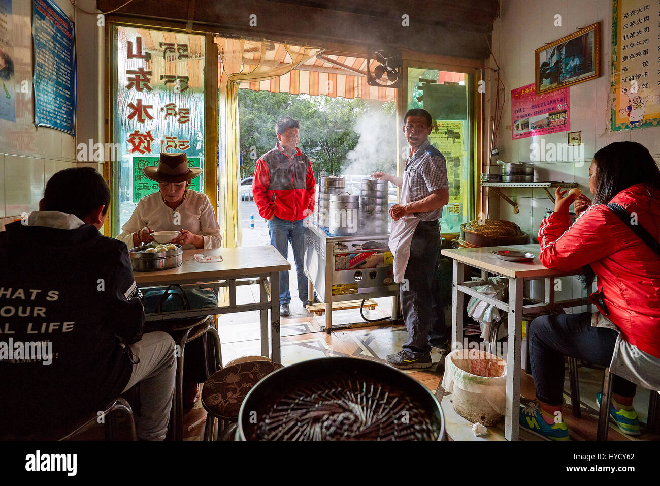 In einem kleinen tibetischen Restaurant in Lhasa, Tibet, essen Gäste dampfende Knödel, während Köche an der hellen offenen Tür mit hohen Dampfern arbeiten Stockfoto