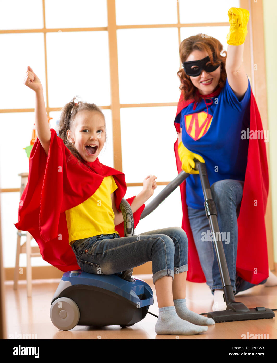 Kind und Mutter verkleidet als Superhelden mit Staubsauger im Zimmer. Familie - Tochter von Frau und Kind viel Spaß während der Reinigung des Bodens. Stockfoto