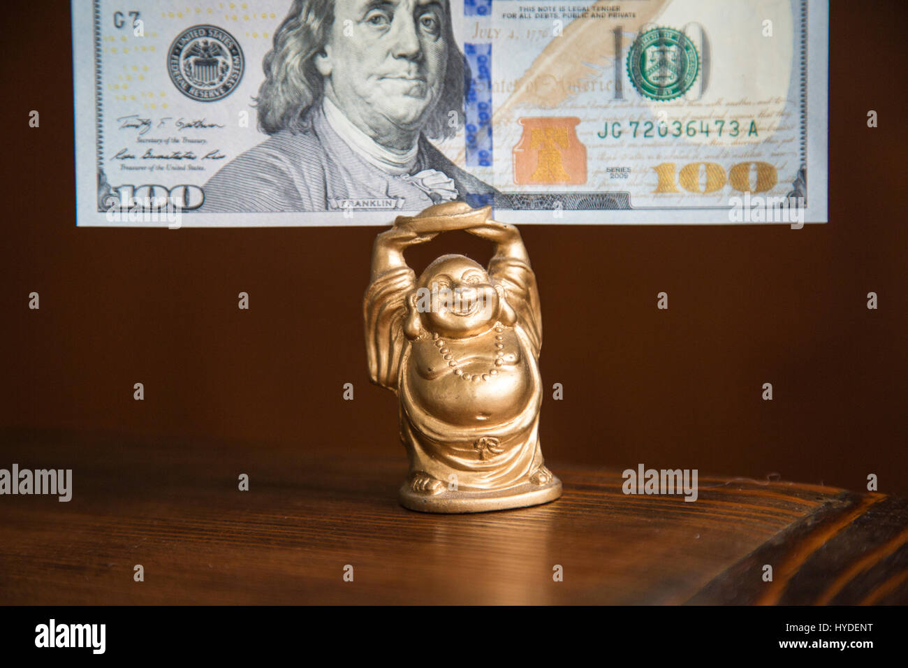 eine kleine goldene Buddha Figur stehend auf einem Holztisch hält einen hundert-Dollar-Schein in Vereinigte Staaten Währung über seinem Kopf Stockfoto