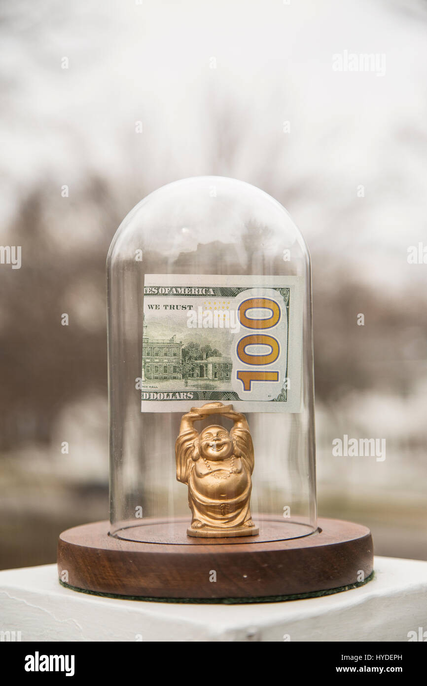 eine kleine goldene Buddha Figur stehend in einem Glas Kuppel hält ein gefalteten hundert Dollar bill über seinem Kopf angezeigt Stockfoto