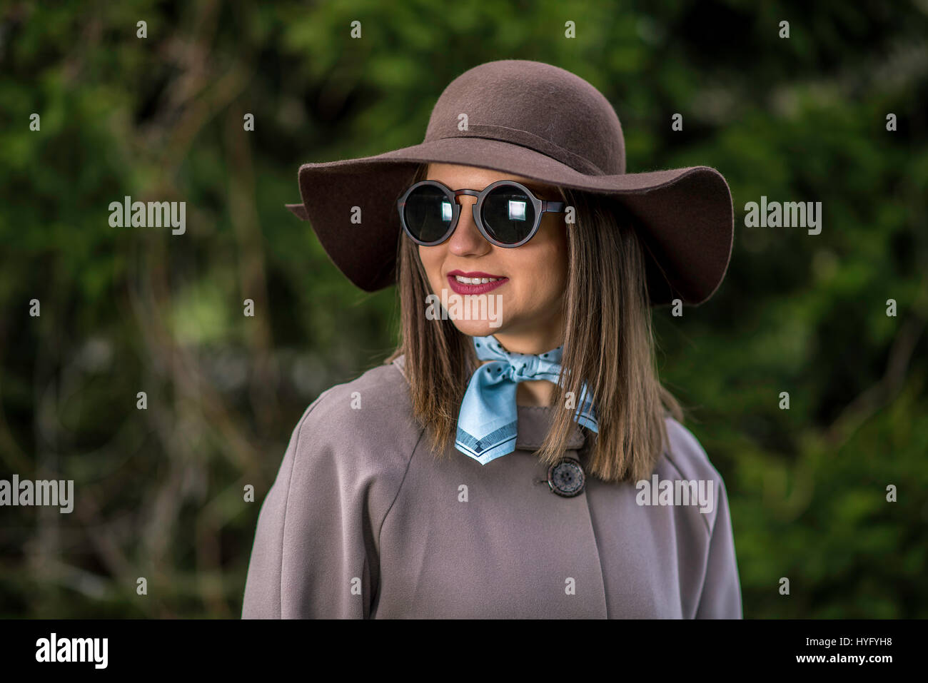 Junge Frau im Regenmantel, Hut und Sonnenbrille posiert im Park bei einem sonnigen Frühlingstag. Geringe Schärfentiefe. Stockfoto