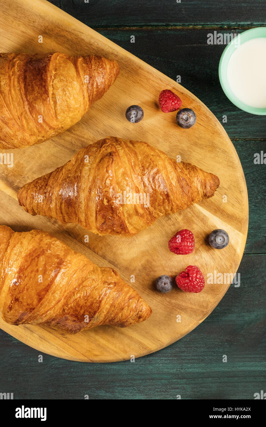 Ein Foto von knusprigen französischen Croissants mit frischen Himbeeren, Heidelbeeren und ein Glas Milch, geschossen von oben auf ein Holzbrett mit einem Platz Stockfoto