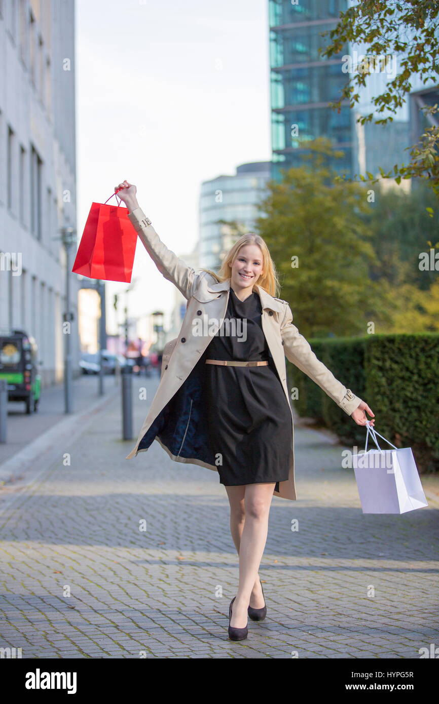 Hübsche blonde Frau mit erhobenen Armen und Einkaufstaschen zu Fuß in der Innenstadt von Stockfoto