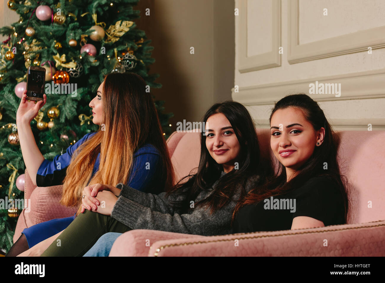 Drei schöne junge Freundinnen machen Selfie auf einem Sofa in der Nähe der Weihnachtsbaum. Horizontale Studioportrait Stockfoto