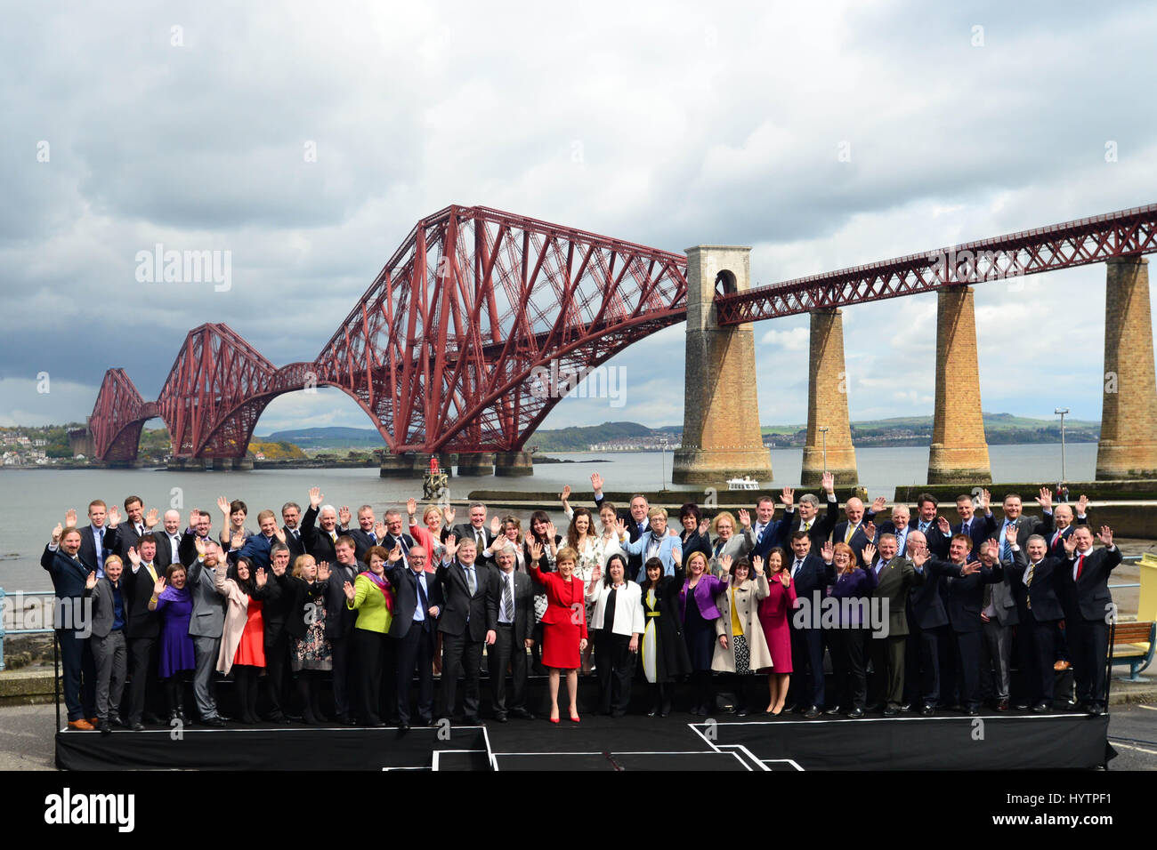 SNP-Führer und schottischen ersten Minister Nicola Sturgeon (C, rot) und die Gruppe der 56 neu gewählte SNP MPs Welle zu den Medien und Fans, als sie für Fotos in South Queensferry mit die Forth Bridge im Hintergrund darstellen Stockfoto