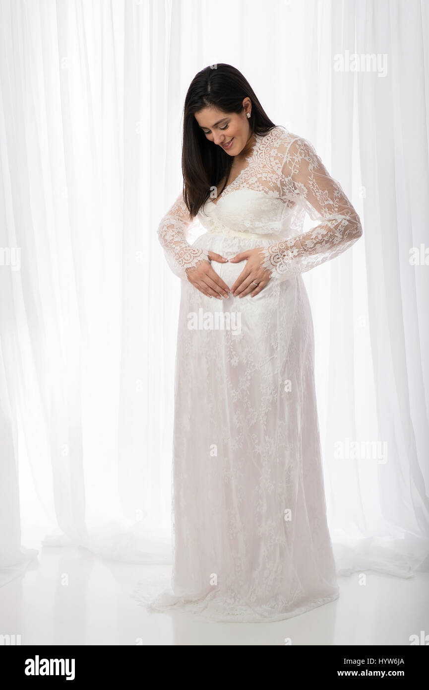 Porträt einer schönen schwangeren Frau trägt eine weiße, Spitze, Ganzkörperansicht, schiere Kleid. Sie lächelt und blickte auf ihren Bauch. Stockfoto