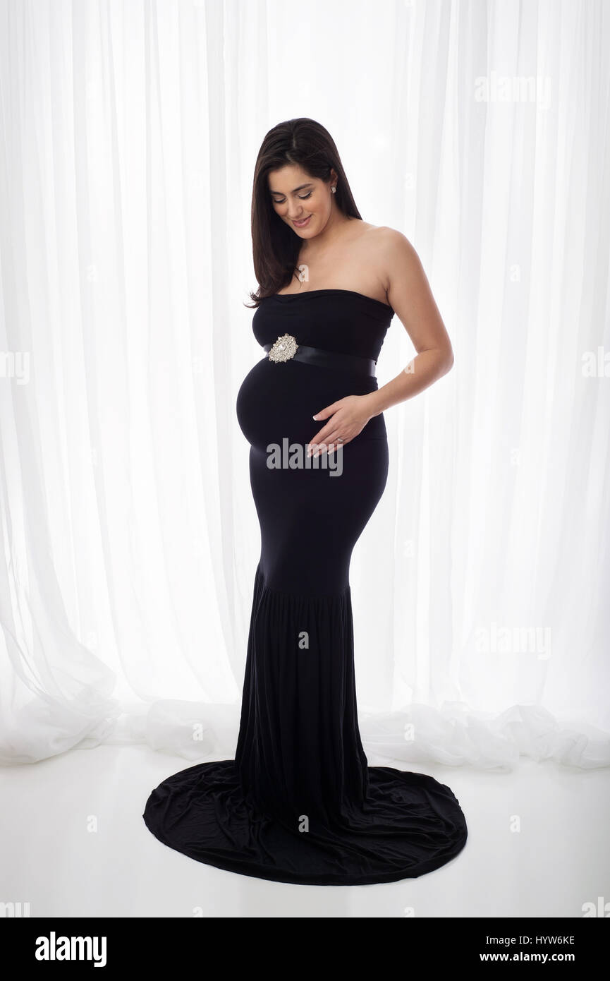 Porträt einer schönen schwangeren Frau trägt eine schwarze, ausgestattet, Meerjungfrau Stil, voller Länge Kleid. Sie lächelt und blickte auf ihren Bauch. Stockfoto