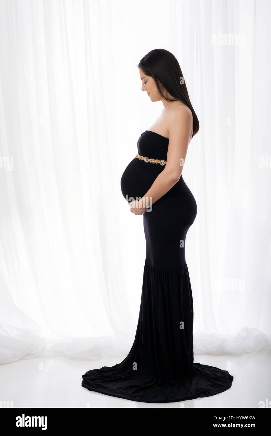 Seite Profilbildnis einer schönen schwangeren Frau trägt eine schwarze, ausgestattet, voller Länge Kleid. Sie lächelt und blickte auf ihren Bauch. Stockfoto