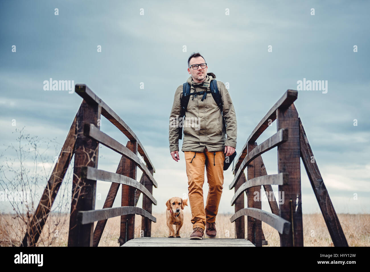 Wanderer und Hund überqueren Holzbrücke am bewölkten Tag Stockfoto
