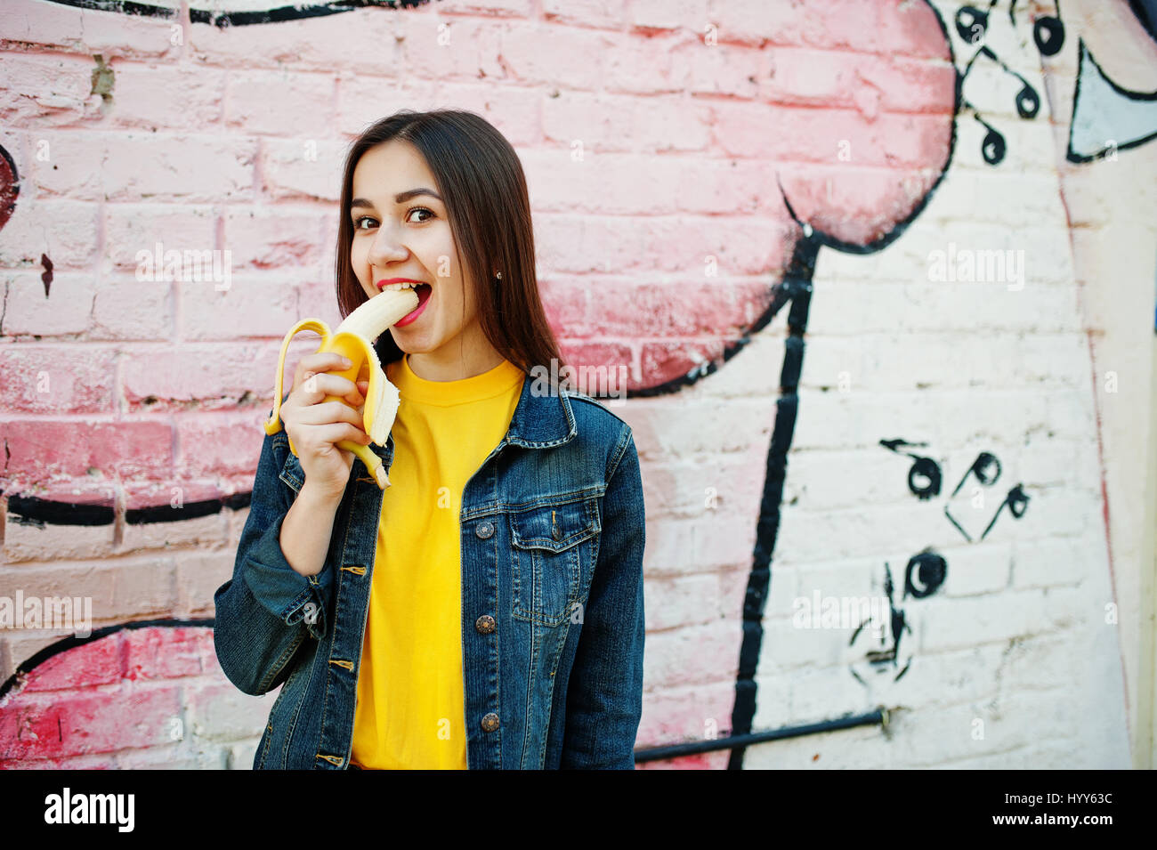 Schöne Teenager-Mädchen essen Bananen, gelbes T-shirt, Jeans in der Nähe von Graffitiwand tragen. Stockfoto