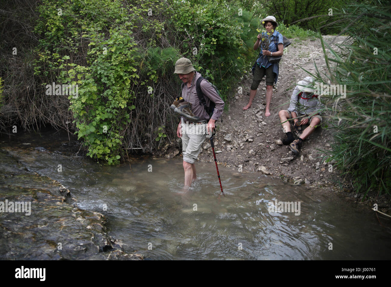 Eine 69 Jahre alte Senioren-Wanderer trägt seine Stiefel während sorgfältig über einen schmalen Bach, seine Freunde ihre Stiefel ausziehen zu bereit, zu folgen. Stockfoto