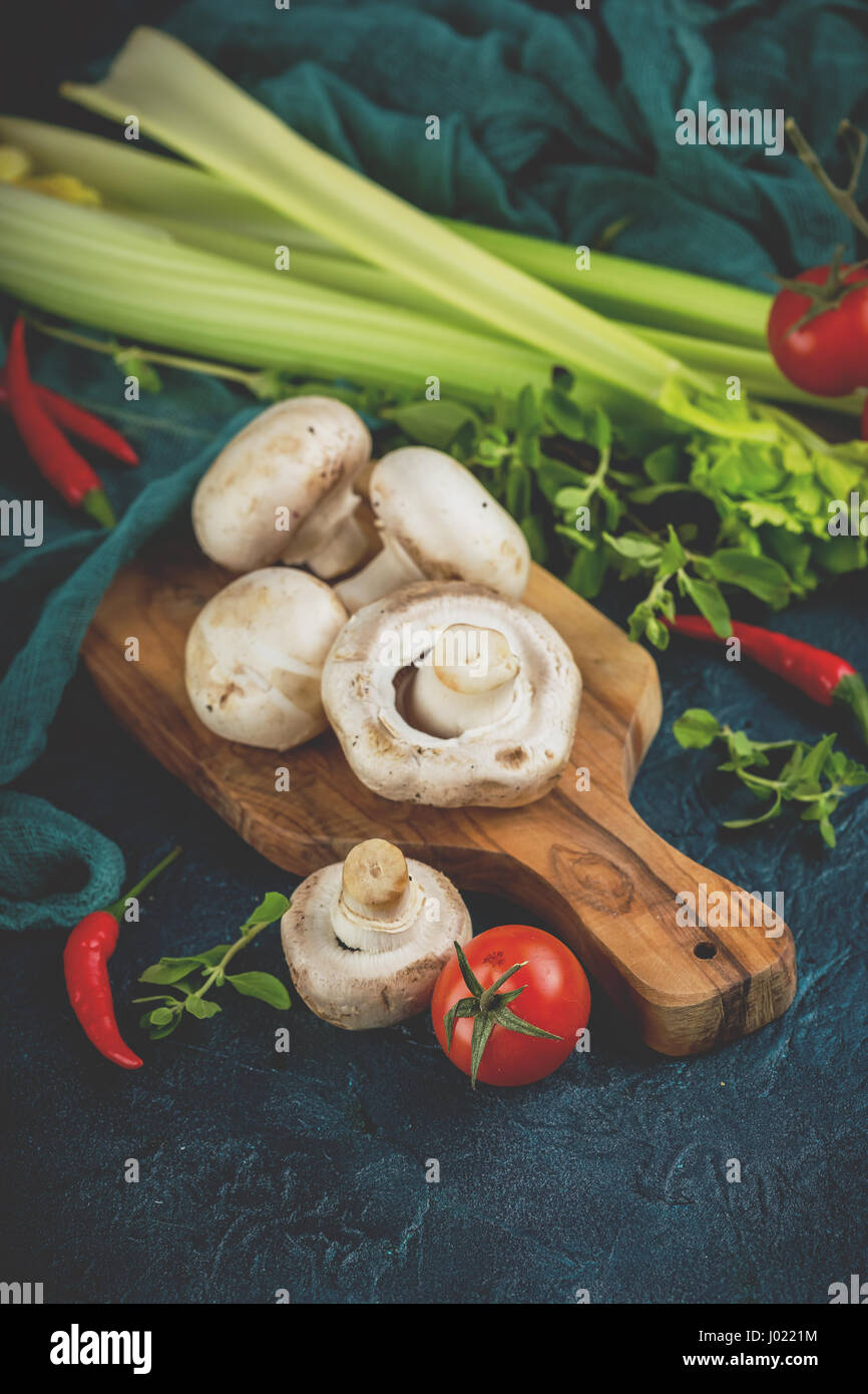 Frischen Champignons Champignons auf ein Holzbrett, Petersilie Grüns, Stangensellerie, Cherry-Tomaten und rote Chilischote auf einem dunklen blau staatlich Stockfoto