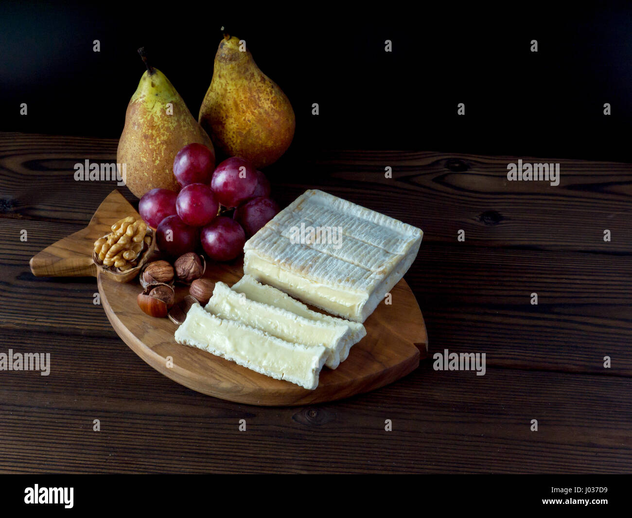 Ziegel geformt Käse mit weißen Schimmel, Haselnüsse, Walnuss, roten Trauben und Birnen auf der strukturierten Holzbrett Stockfoto