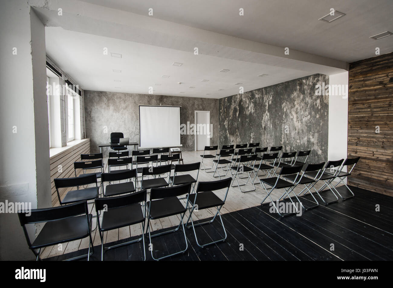Raum für Vortrag mit vielen dunklen Stühlen. Wände sind weiß, Loft Interieur. Auf der rechten Seite ist eine Tür. Auf dem Hintergrund gibt es einen Tisch mit einem Laptop. Stockfoto