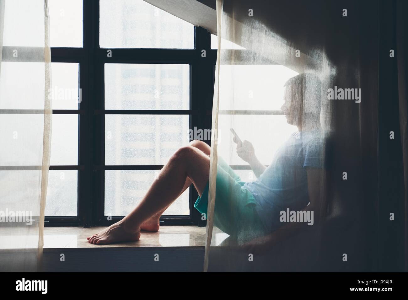Einsamer trauriger Mann hinter dem Vorhang mit Handy am Fenster sitzen, während regnerischen Tag. Stockfoto