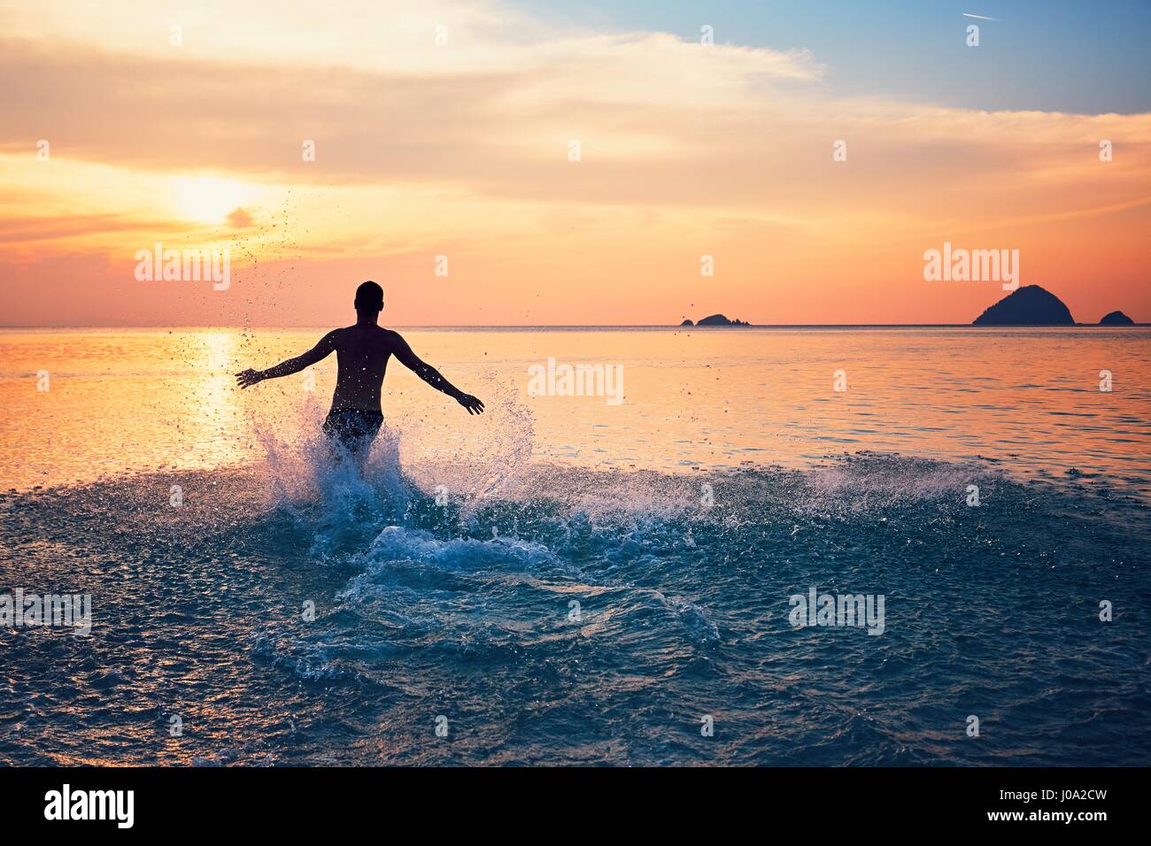 Freudiger junger Mann läuft in das tropische Meer bei Sonnenuntergang. Stockfoto