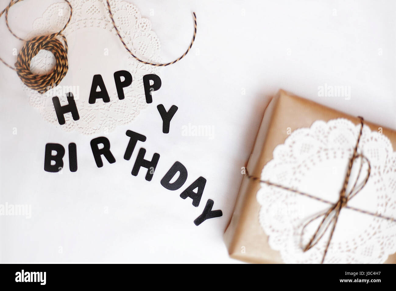 Geschenk verpackt in braunem Papier, Deckchen und Zeichenfolge neben Buchstaben Happy Birthday Stockfoto