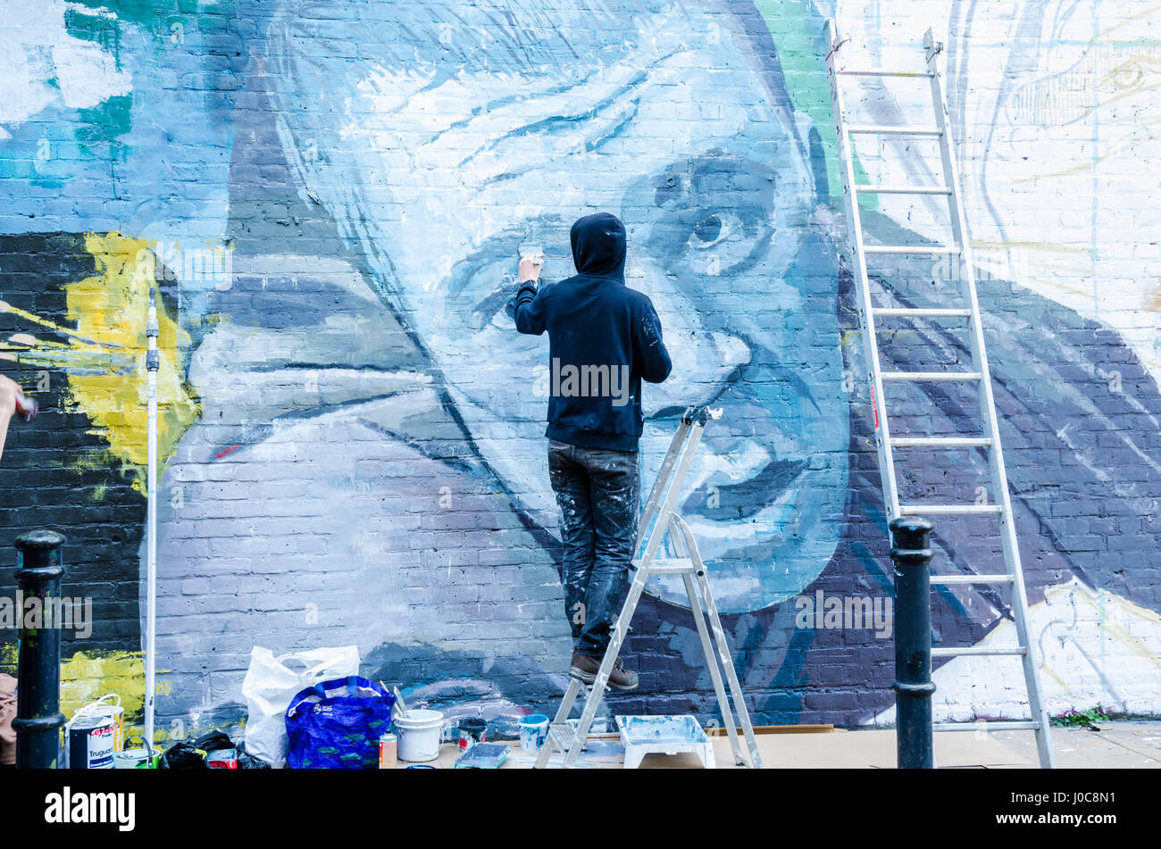 Ein Künstler, trug eine schwarze Kapuzenjacke schafft ein Kunstwerk durch das Malen ein Wandbild an der Wand in der Hanbury Street in East London. Stockfoto