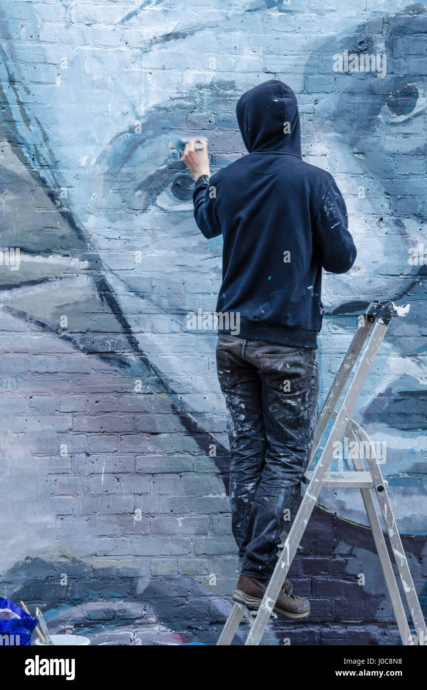 Ein Künstler, trug eine schwarze Kapuzenjacke schafft ein Kunstwerk durch das Malen ein Wandbild an der Wand in der Hanbury Street in East London. Stockfoto