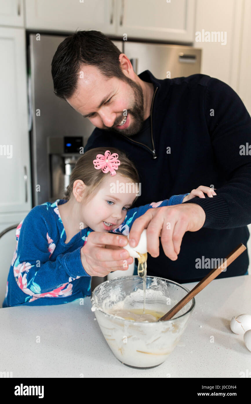 Mitte erwachsener Mann und Tochter knacken Ei in Rührschüssel geben gemeinsam am Küchentisch Stockfoto
