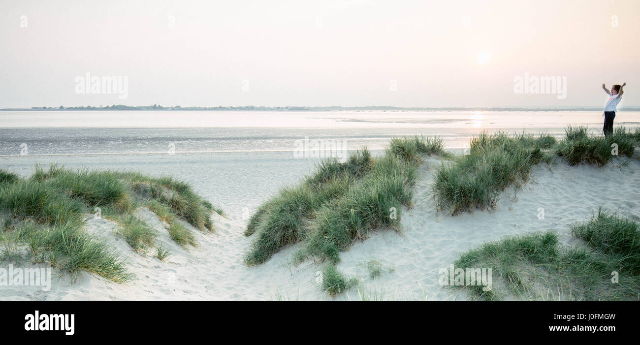 Ein kleiner Junge wirft seine Arme in der Luft in Freude, an einem Sandstrand am Sonnenuntergang spielen unter den Sanddünen zu sein. Stockfoto