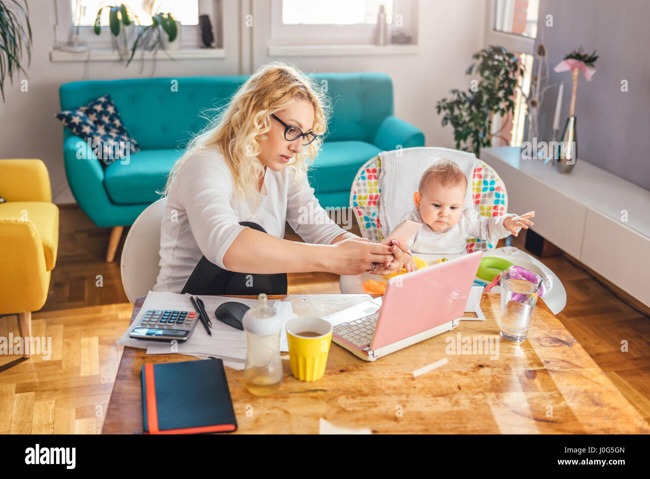 Mutter trägt Brille mit ihrem Baby am Schreibtisch sitzt und mit Smartphone zu Hause im Büro Stockfoto