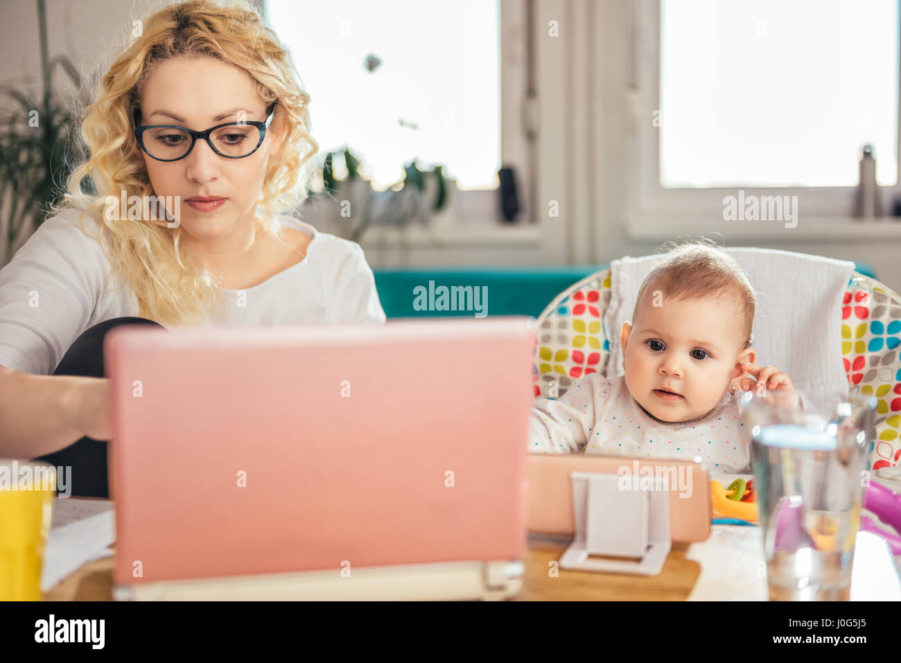 Mutter Tragen von Brillen am Schreibtisch sitzen und arbeiten am Laptop während Ihr Baby cartoon beobachten auf dem Smartphone Stockfoto