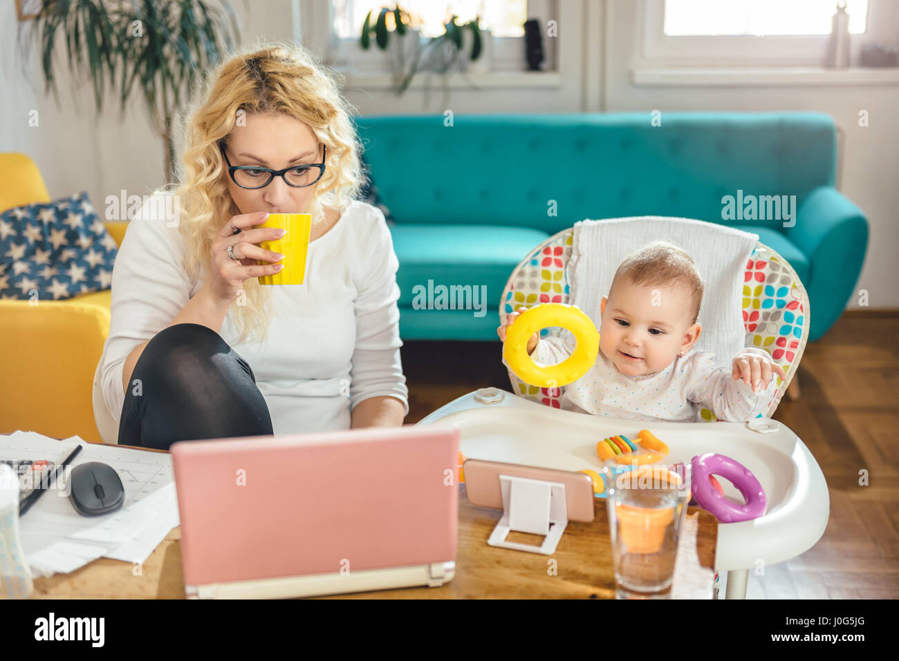 Mutter Tragen von Brillen an einem Schreibtisch sitzen, Kaffee trinken und Arbeiten am Laptop während Ihr Baby cartoon beobachten auf dem Smartphone Stockfoto