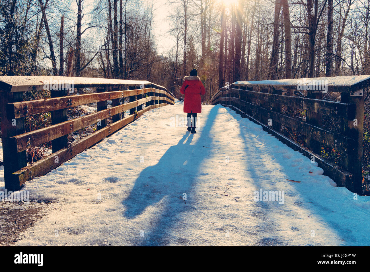 Frau im roten Mantel werfen lange Schatten über Schnee bedeckte Brücke im Wald wandern. Stockfoto