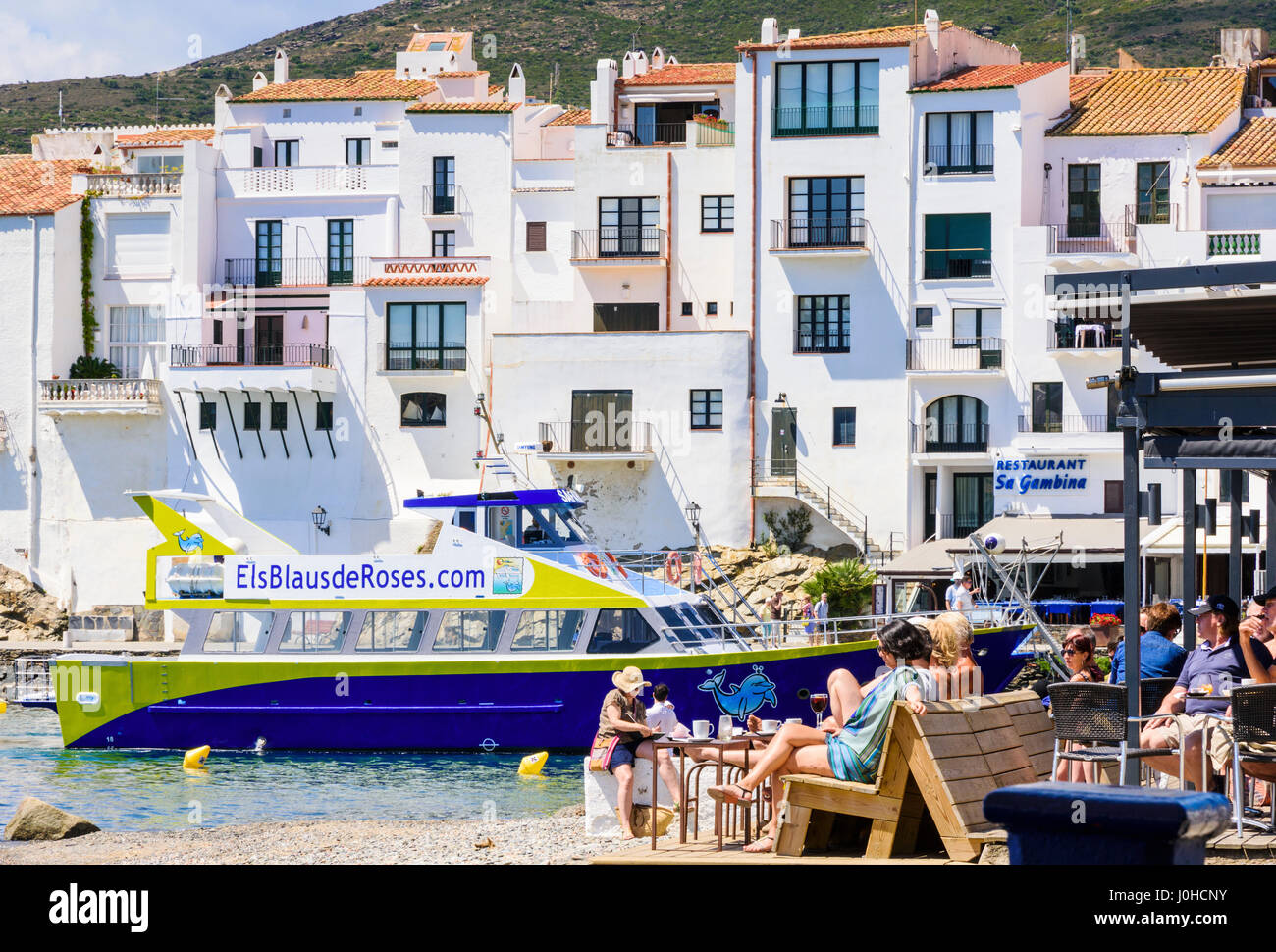 Beliebte Waterfront Beachbar am Hauptstrand Cadaques, überragt von den weiß getünchten Stadt und vertäut Touristenboot, Cadaques, Katalonien, Spanien Stockfoto