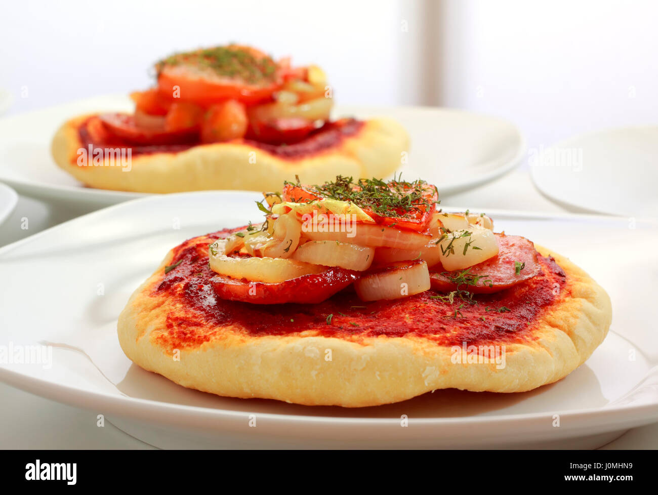 Kleine Pizza (Pizzette) auf Teller mit Wurst, Zwiebeln und Tomaten Scheibe. Unkonzentriert zweite Pizza auf Hintergrund. Stockfoto