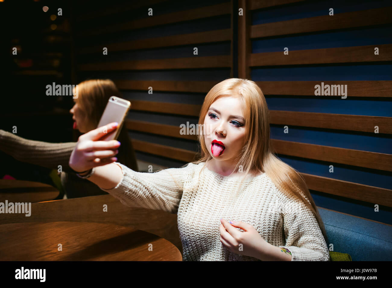 Junge, blonde Frau in einem Strickpullover, Zunge, sitzen an einem Tisch in einem Café, verbringen Zeit al zeigt, ein Selfie Foto auf einem Smartphone emotional Stockfoto