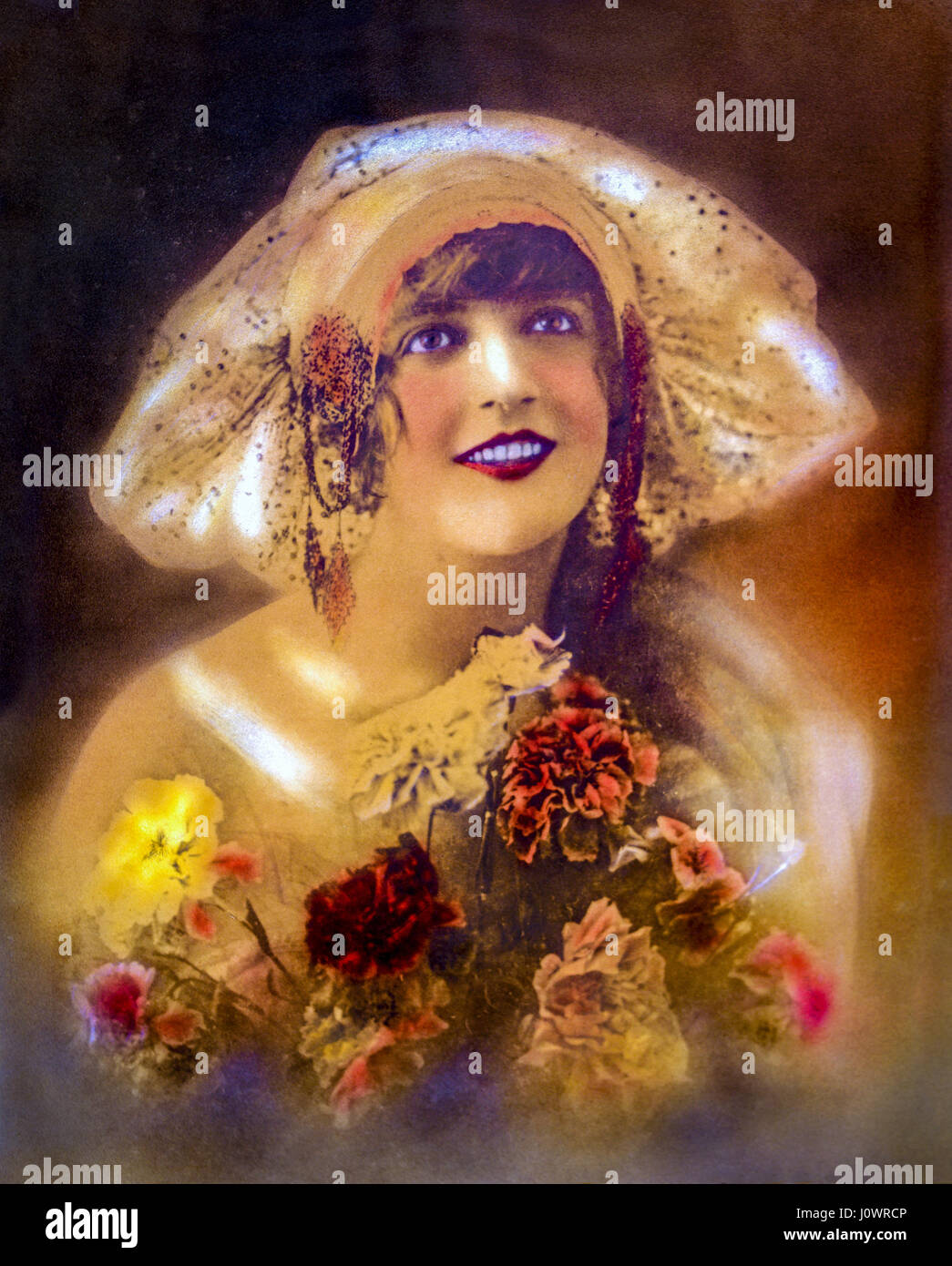 Handkolorierten Vintage Porträt einer lächelnden 1920er Jahre Ära Flapper Mädchen in ihren Zwanzigern, mit breiter Krempe Cloche Hut mit einem Bouquet von Nelke Blume Stockfoto