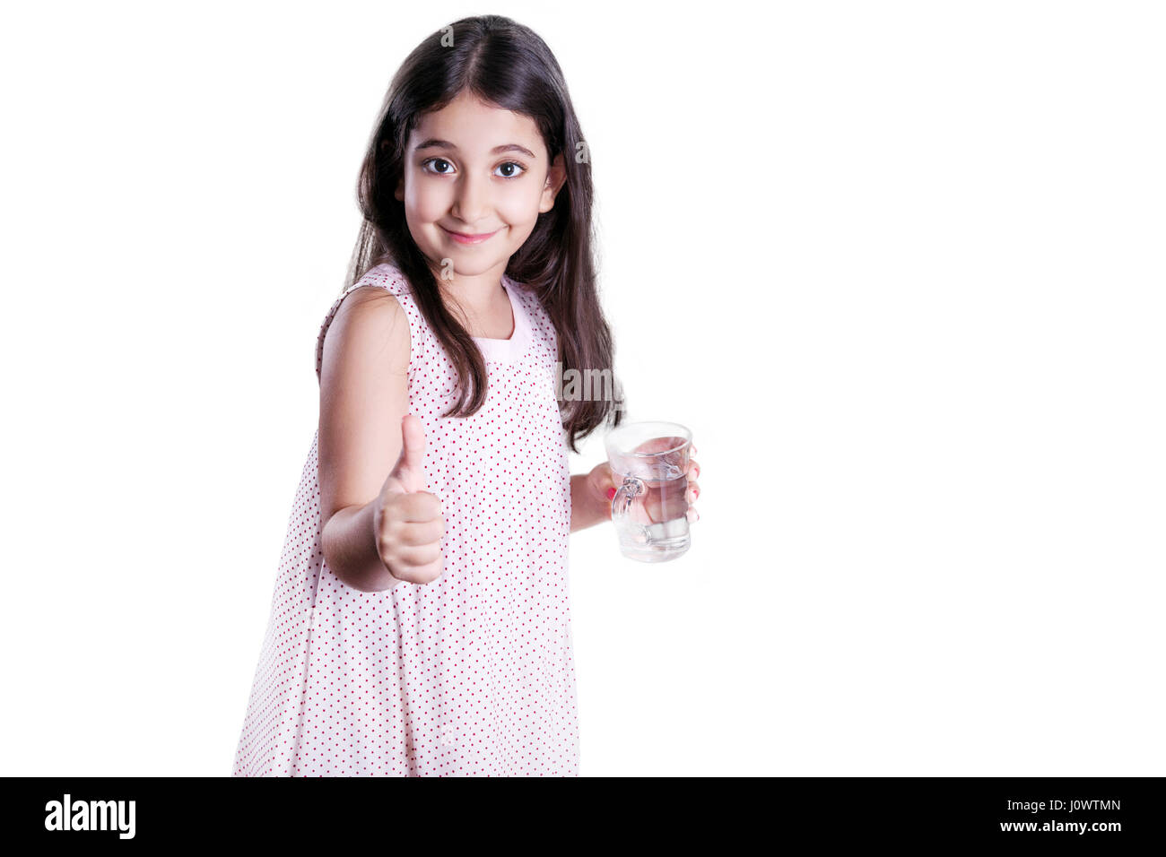 Schöne glückliche kleine Mädchen mit lange dunkle Haare und Kleid Glas Wasser hält und zeigt Daumen. Studioaufnahme, isoliert auf weißem Hintergrund. Stockfoto