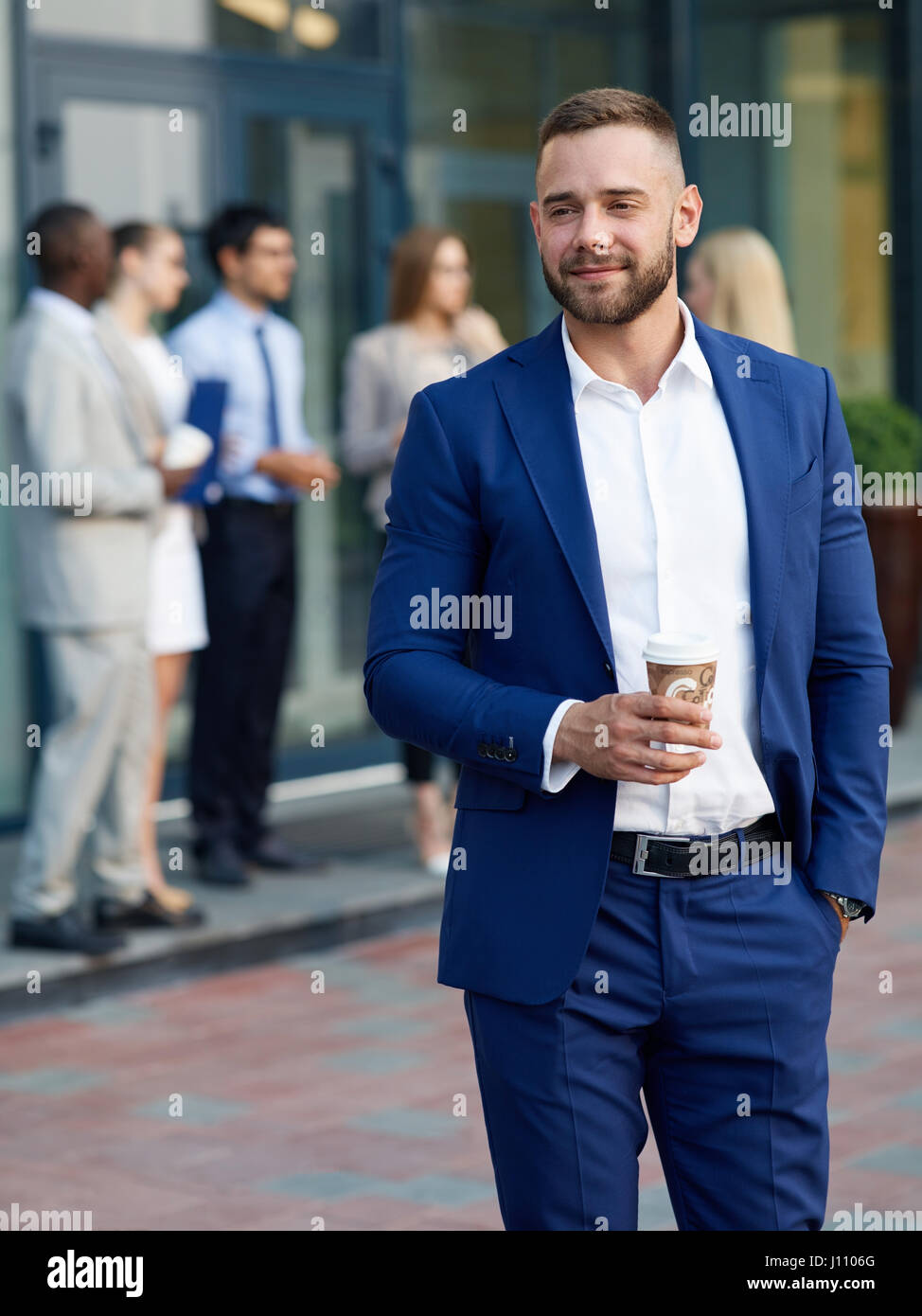 Porträt von schönen jungen Mann tragen stilvolle blau passen lächelnd und trinken Kaffee aus Pappbecher außerhalb Bürogebäude, Gruppe von Personen in backgr Stockfoto
