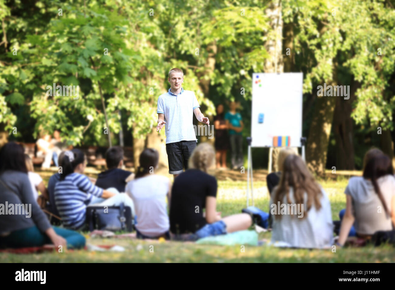 Eine Gruppe von jungen Studenten in den Park. Ansicht eines Mannes gestikulieren mit den Händen gegen einen defokussierten Gruppe von Menschen, die vor ihm saß auf dem Rasen stehen. Stockfoto