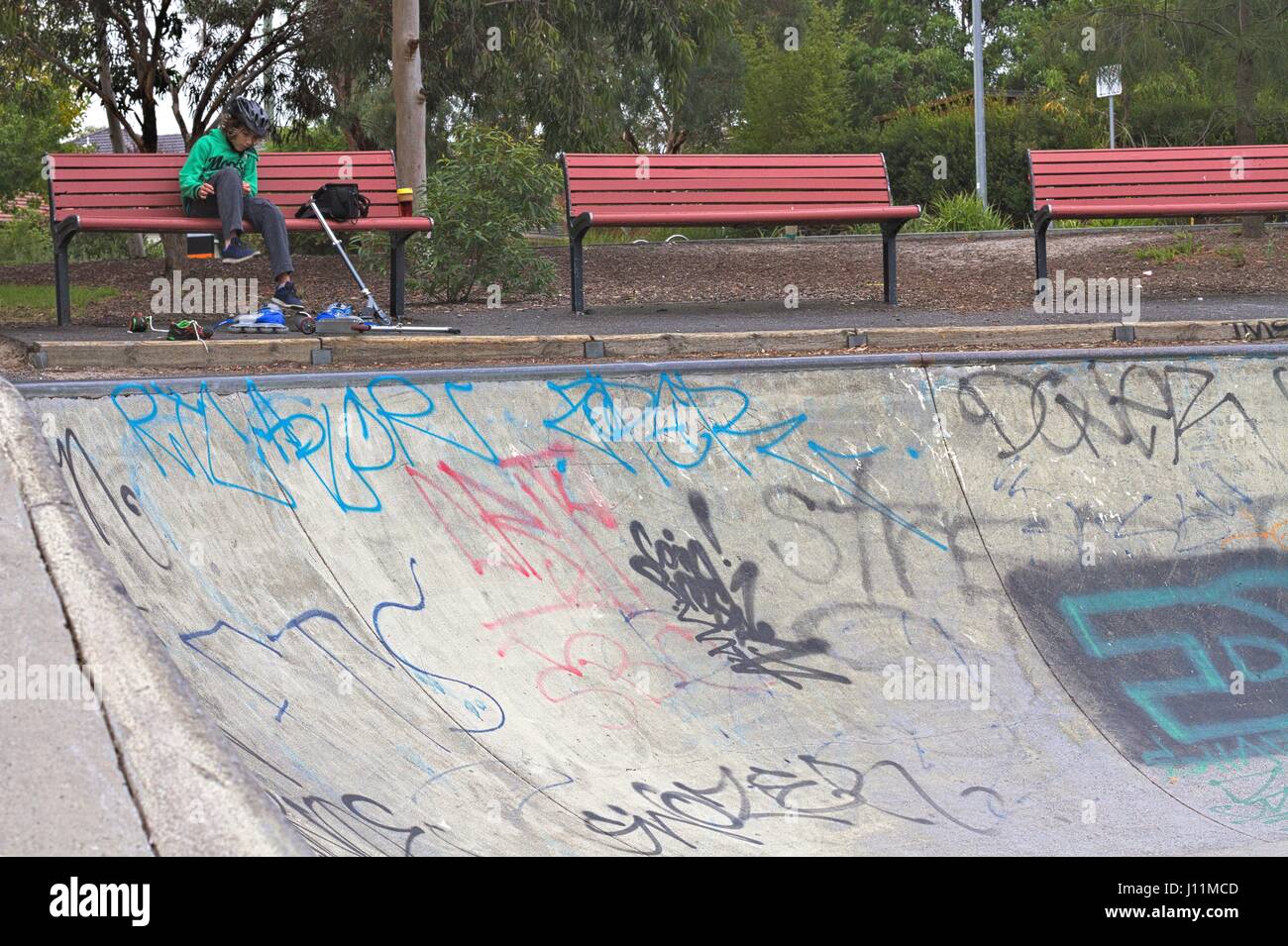 Drei rote Sitzbänke und ein Junge mit einem Scooter mit Blick auf ein Graffiti bedeckt half-Pipe in einem Skatepark. Stockfoto Drei rote Sitzbänke und ein Junge mit einem Scooter mit Blick auf ein Graffiti bedeckt half-Pipe in einem Skatepark. Stockfoto