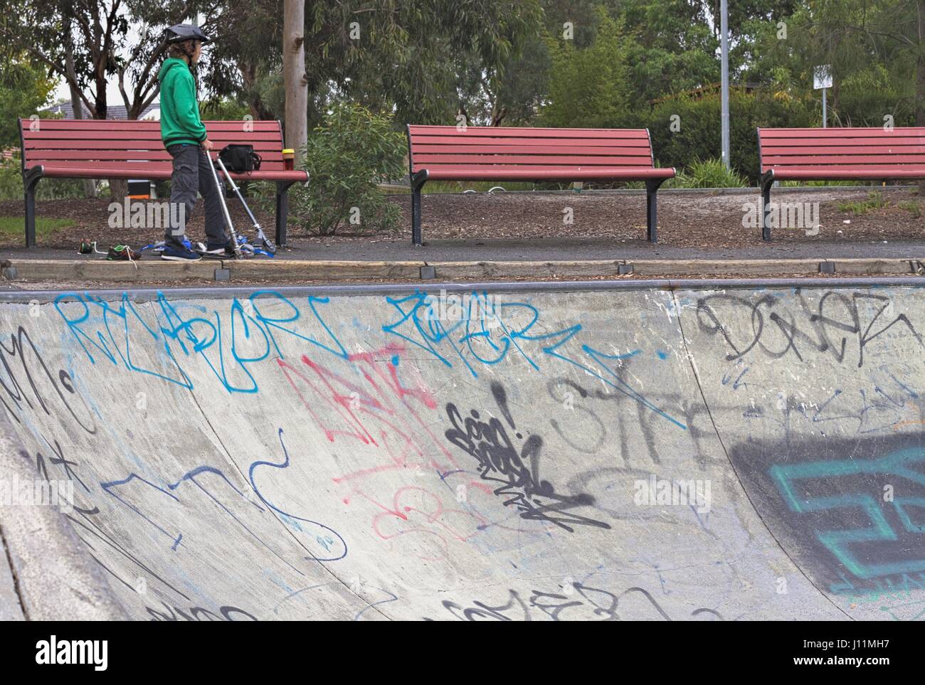 Drei rote Sitzbänke und ein Junge auf einem Roller mit Blick auf ein Graffiti bedeckt half-Pipe in einem Skatepark. Stockfoto Drei rote Sitzbänke und ein Junge auf einem Roller mit Blick auf ein Graffiti bedeckt half-Pipe in einem Skatepark. Stockfoto