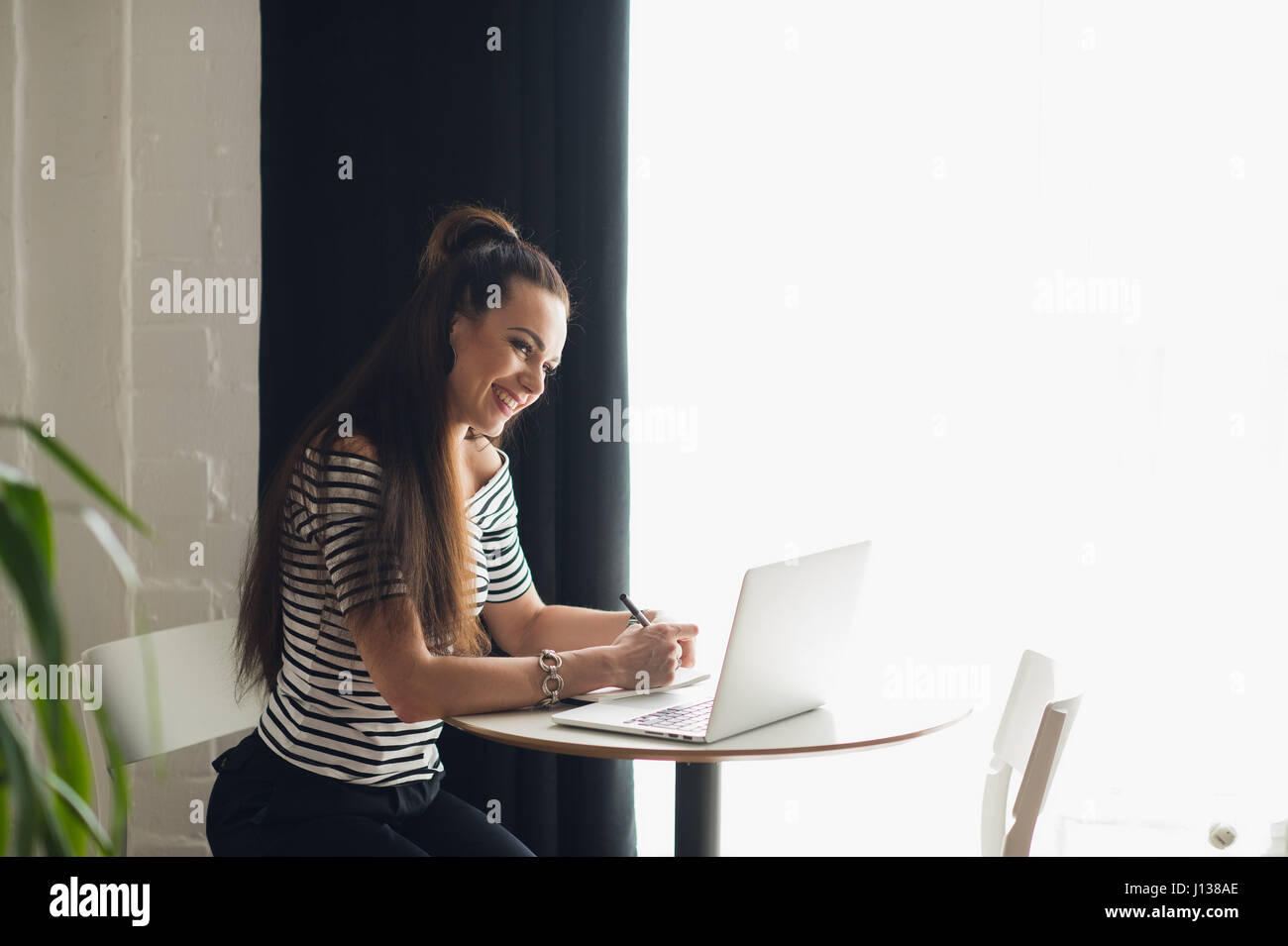 Foto von hübsche Frau Notizen machen und denken über ein Thema während der Sitzung in einigen Coffee-Shop mit ihrem Laptop in der Nähe großer Fenster. Stockfoto