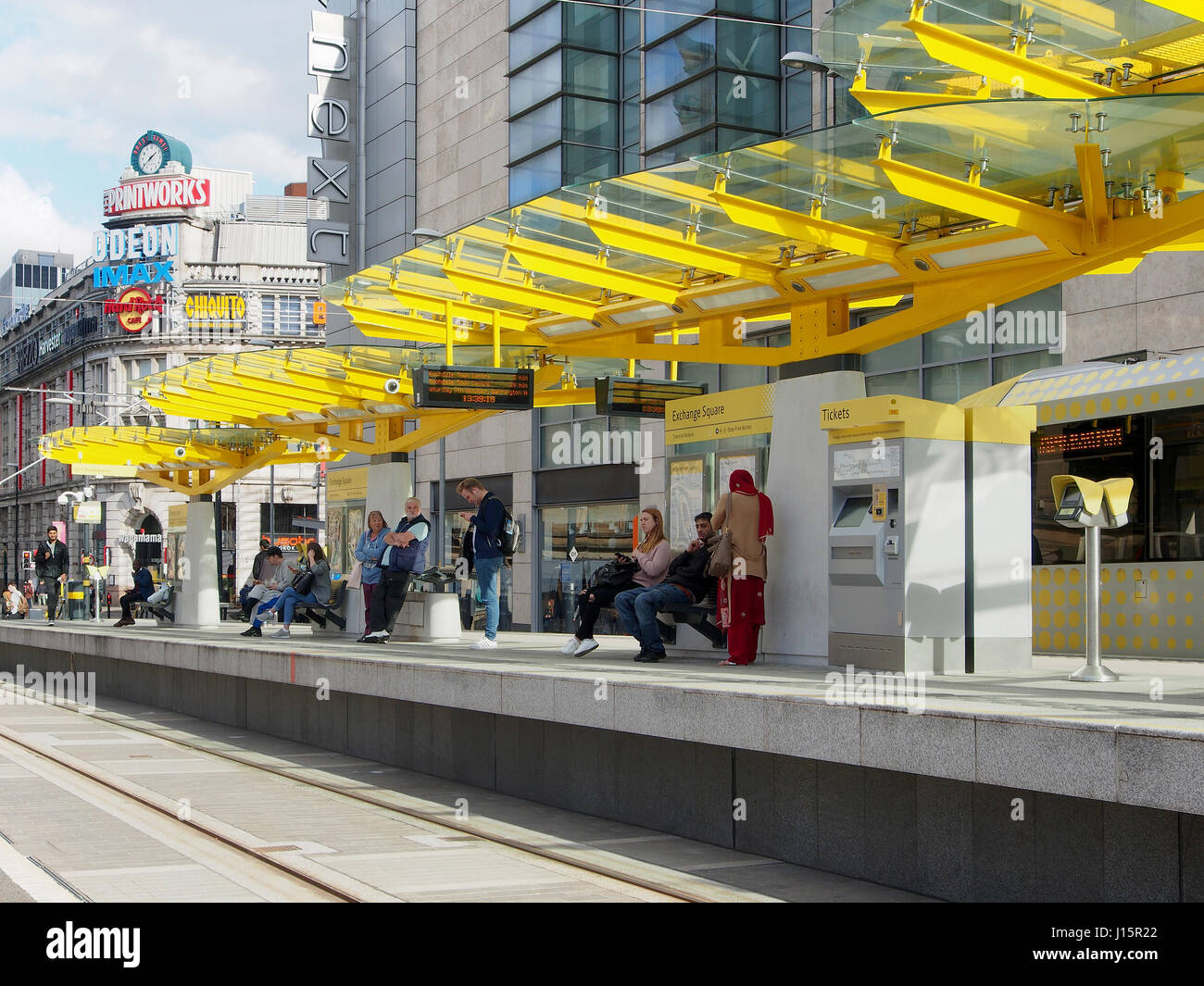 Exchange Square im Zentrum von Manchester, England, zeigt Menschen auf der Straßenbahn-Plattform, mit der bunten farbenfrohen Printworks im Hintergrund. Stockfoto